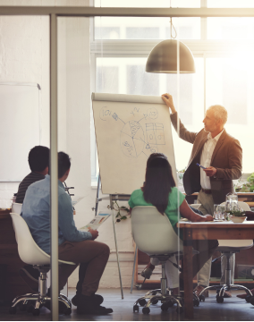 Um homem mais velho de terno bege apresenta um gráfico desenhado em um flip chart para um grupo de três pessoas sentadas ao redor de uma mesa em um ambiente de escritório moderno e iluminado.