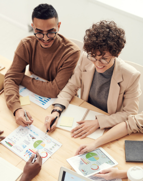 Duas pessoas sorridentes, um homem e uma mulher, analisam gráficos e estatísticas impressos em papéis e exibidos em um tablet, enquanto fazem anotações em um ambiente de escritório.