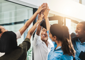 Grupo de pessoas em um ambiente corporativo celebrando com um toque coletivo de mãos no alto, sorrindo e demonstrando entusiasmo.